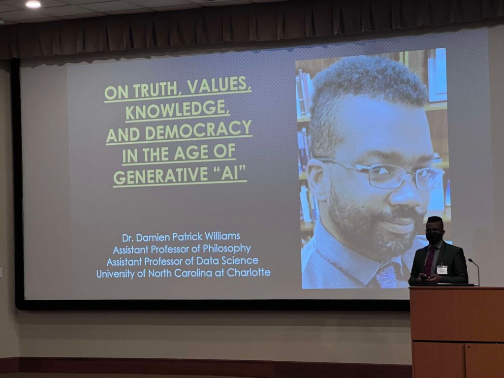 A Black man with natural hair shaved on the sides & long in the center, grey square-frame glasses, wearing a silver grey suit jacket, a grey dress shirt with a red and black Paisley tie, and a black N95 medical mask stands on a stage behind a lectern and in front of a large screen showing a slide containing the words On Truth, Values, Knowledge,and Democracy in the Age of Generative “AI”Dr. Damien Patrick Williams Assistant Professor of Philosophy Assistant Professor of Data Science University of North Carolina at Charlotte, and an image of the same man, unmasked, with a beard, wearing a silver-grey pinstriped waistcoat & a dark grey shirt w/ a purple paisley tie in which bookshelves filled w/ books & framed degrees are visible in the background